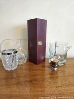 Full lot displaying all items on a wooden surface against a white wall. Items include wooden cocktail tool case, miniature champagne bucket with toothpicks, cut crystal decanter, cut glass ice bucket with chrome handle and tongs, Krosno crystal ice bucket with tongs.