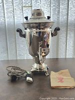 Front view of the electrosamovar tea pot. Polished metal body with curved handles on either side, spigot in front, electric cord and plug coiled beside it, paper manual or instructions nearby.