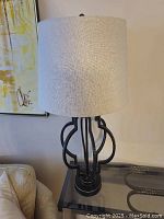 Full view of the table lamp showing geometric metal base and natural linen shade on glass table near wall with artwork and cream couch.