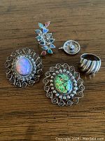 Five pieces of vintage silver jewelry on wooden surface showing two filigree brooches with colorful glass cabochons, two rings, and one hat charm.