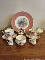 Overview image of all items in the lot including decorative plates, teacups with saucers, small pitchers, floral bouquet, trinket box, and jam pot.