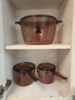 Large smoked Pyrex pot on upper shelf, two smaller saucepans with handles below inside kitchen cabinet