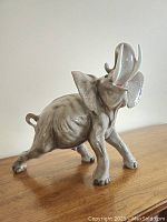 Front-left perspective view of the ceramic elephant sculpture on wooden surface, showing the full body and raised trunk.