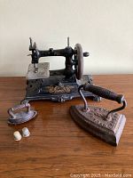 Cast iron Muller toy sewing machine with floral design, cast iron iron, miniature iron model, and two vintage metal thimbles on wooden surface.