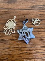 Three vintage Judaica pendants laid on wooden surface: gold hand pendant with circular dove medal, silver Star of David with Chai pendant, small 14K gold Chai charm