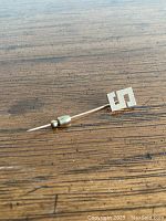 Stickpin laid flat on wood surface showing block letter S head at one end and metal fastener at the other.