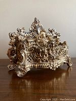 Front angled view showing ornate scrollwork, lattice, gadroon details, and bowed front compartment on gilt cast metal letter rack.