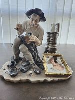 Full front view of the shoemaker figurine seated, holding a shoe patch and needle.