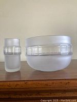 Two frosted glass pieces: a bowl and a vase with clear geometric bands, set on wood surface against a plain background
