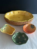 Large yellow floral nesting baking dish with woven base, three vegetable shaped serving dishes in front: yellow pepper, orange pumpkin, green artichoke.