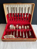 Overall view of the flatware set in the box showing the knives, forks, spoons, and serving pieces with the box interior lined in red velvet and cream satin fabric. Wooden box has some wear and broken hinges.