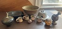 Wide shot of all pottery items on table including oil lamp, bowl, trivet, mugs, bell, mortar and pestle, spherical object