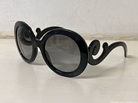 Black Prada sunglasses with large round dark lenses and ornate curvy arms shown on side view. Background white surface and wall.