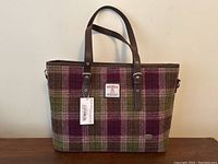 Frontal view of the tote bag showing plaid Harris Tweed wool exterior, brown handles and attached tags
