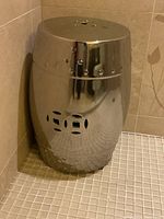 View of the stainless steel garden stool positioned in a corner shower area, showing glossy reflective surface and cut-out square pattern on the top.