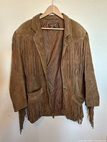 Full front view of vintage brown suede leather fringe jacket on hanger showing shoulder and pocket fringes and interior lining.