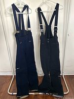 Two pairs of navy ski pants with suspenders hanging side by side, showing full front view