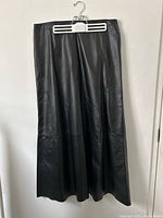 Full length view of black leather skirt on white plastic hanger against white wall showing skirt texture and length.