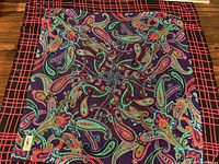 Full view of scarf showing colorful paisley design and black border with red grids on wooden surface.