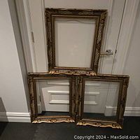 Three large wooden picture frames with carved decorative bronze finish leaning against a white wall. One frame is at the back, two in front.