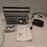 Photo showing all three items: Sony digital camera with attached wrist strap, Panasonic AM/FM radio, Westclox digital alarm clock, and Sony user manual.