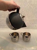 Photo showing polished stainless steel and black accent Matilda coffee maker held in hand with two matching small polished stainless steel espresso cups on white surface.