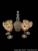 Six amber hued wine glasses arranged in a semicircle around a clear glass decanter with stopper on a black background.