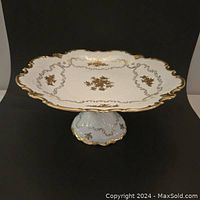 Front angled view of white porcelain cake stand showing square top with gold floral patterns and scalloped edges with gold trim.