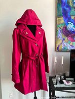 Front view of women's red Calvin Klein trench coat with hood on dress form, showing button closure and waist belt.