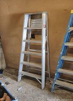 Full view of the 5-step aluminum A-frame ladder leaning against a wall indoors, showing both front and side steps.