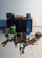 Photo showing the black metal tackle box closed, with fishing lures, hooks, lines, and other fishing gear arranged around it on a white cloth.