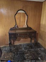 Front view of dark wooden antique dressing table with attached ornate mirror. Displayed in a corner against wood panel walls.