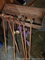 Image showing six wooden croquet mallets standing with visible wear and patina, multiple croquet balls wrapped in purple cloth, and a long rectangular wooden storage box with aged wood and surface wear.