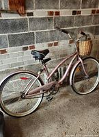 Full side view of the pink ladies Sport Tech bicycle leaning against a brick and concrete wall. Shows overall frame, tires, seat, and basket.