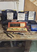Wide view of the boxed tool sets and scattered hand tools on a workbench including ratchet socket set, manuals, and pliers