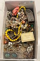 Overhead photo of a box filled with assorted vintage costume jewellery pieces and loose parts, including beaded necklaces, bracelets, and brooches.