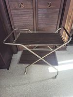 Full view of mid-century modern style bar cart showing two tiers of faux wood grain shelves and brass tone frame.