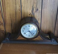 Front view of wooden mantle clock with rounded top housing clock face, decorative inlay on front, and winding key placed beside clock on surface