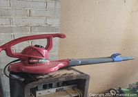 Red Toro electric blower vac with large handle and extended black nozzle with blue tip, shown resting on a black metal cabinet.