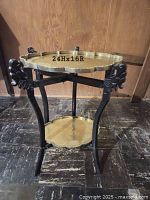 Full view of the two-tiered ebonized carved wood tray stand with brass-colored scalloped edge trays on floor against wood-paneled wall.