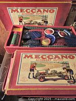 Open red Meccano box showing metal parts including wheels and strips inside with illustrated box lid.