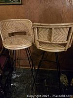 Pair of vintage rattan barstools with woven backs and seats on black metal hairpin legs, placed against a wooden background.