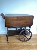 Side view of the folded tea cart showing the wooden fold down sides and one large rear wheel with two front caster wheels.