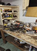 Wide shot of a wooden workbench cluttered with assorted tools, hardware parts, tapes, clamps, hand tools, and containers. Shelves behind contain more small items and containers.