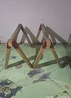 Pair of wooden workhorses set up in a concrete floor basement area. Each workhorse consists of 4 angled legs connected with a flat wooden beam, reinforced with red metal brackets on each joint.