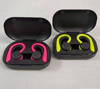 Two pairs of Bluetooth sport earbuds in open black charging cases, one with pink ear hooks, the other with neon yellow-green ear hooks, shown side by side.