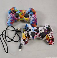 Two wireless PS3 game controllers with colorful splatter paint designs and one USB charging cable placed on a gray surface.