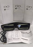 One pair of black 3D active glasses with USB charging cable and user manual laid out in front of three boxes.