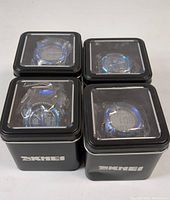 Four SKMEI digital watches in black metal display tins, showing watch faces through clear lids arranged in grid.