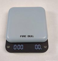Top view of the light blue Fire Bull digital coffee scale showing black display panel with digital timer and weight readout, touch buttons labeled POWER TIMER, g/oz/ml and ZERO.